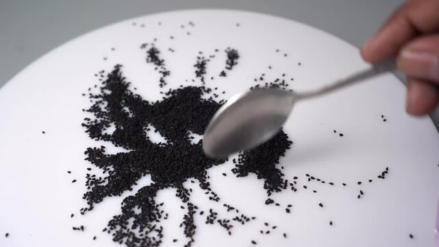 Close-up view of hand use spoon to form black sesame seeds on white plate, create abstract art design pattern with organic ingredient