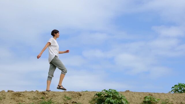 Young brunette woman walks along gravel road past lush green bushes and blooming summer wildflowers. Peaceful rural landscape of Croatia offers relaxation and slow living vibes.