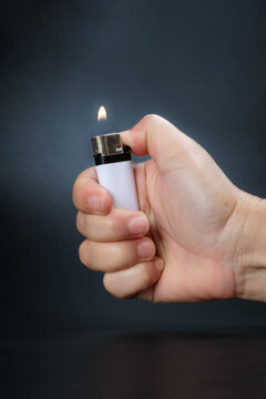 Close-up of a person's hand igniting a white disposable plastic lighter with a small flame against a dark background.