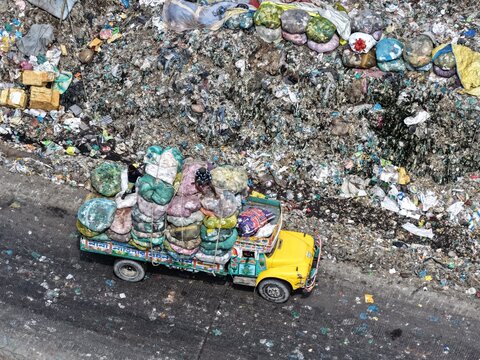 Aerial view of a yellow truck heavily loaded with large sacks of waste driving past a massive landfill site in Dhaka, Dhaka Division, Bangladesh.