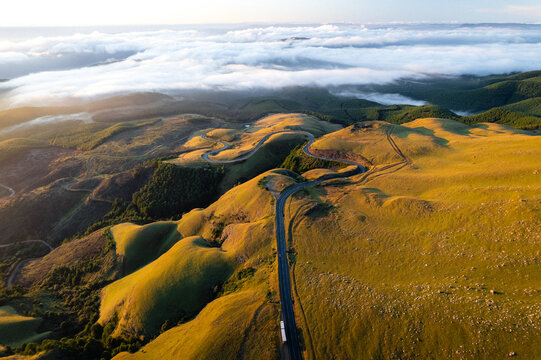 Aerial view of Long Tom Pass winding through the rolling green hills of Mpumalanga during golden hour with clouds in the distance in Ehlanzeni District Municipality, Mpumalanga, South Africa.