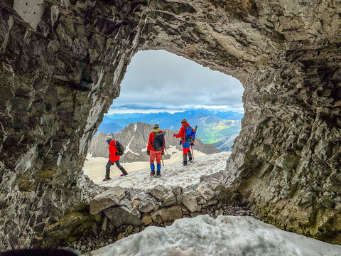 Hikers in bright outdoor clothing framed by a natural rock opening above lingering snow in the high mountains of the French Pyrenees.