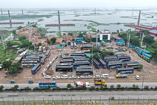 Aerial view of a busy bus depot with numerous parked buses and cars, adjacent to a flooded area with brick kilns in Dhaka, Dhaka Division, Bangladesh.