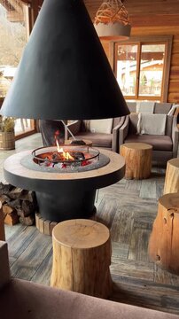A cozy indoor setting featuring a modern fireplace with a conical hood. Surrounding wooden stools and comfortable chairs create a warm atmosphere.