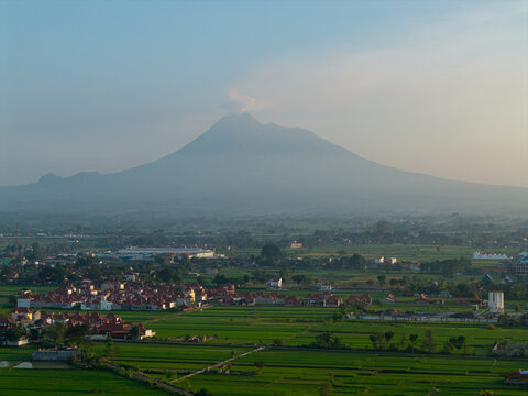 Aerial view of Mount Merapi volcano with smoke rising above lush green rice fields and residential areas in Kecamatan Prambanan, Jawa Tengah, Indonesia.