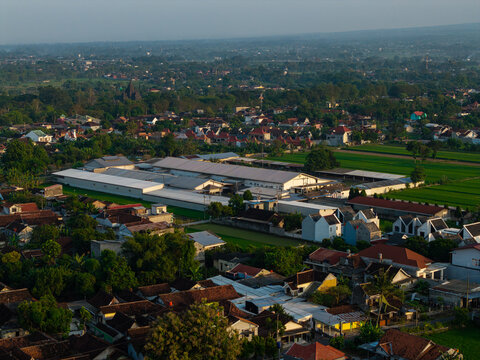 Aerial view of a large industrial facility surrounded by residential houses, lush green rice fields, and trees in the rural landscape of Kecamatan Prambanan, Jawa Tengah, Indonesia.
