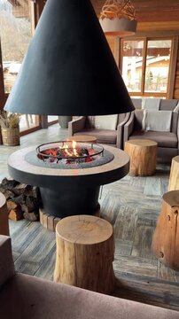 A modern interior featuring a circular fireplace with a black chimney. Surrounding are wooden stools and a cozy seating area with gray chairs.