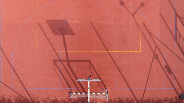 Aerial view of a basketball court with a red surface, yellow court markings, and long shadows cast by the hoop and surrounding structures in Gdansk, Poland.