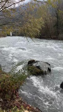 A flowing river with rocky banks and autumn foliage. The water is turbulent, creating whitecaps. Trees with yellow leaves line the riverbank.