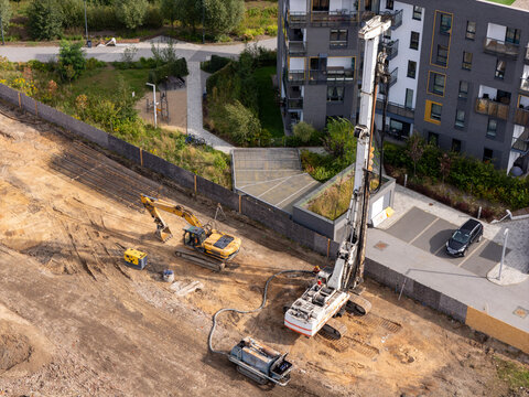 Aerial view of a construction site with an excavator and a drilling rig operating on a dirt lot next to modern residential buildings in Gdansk, Poland.