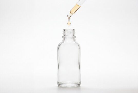 A glass dropper dispensing a golden liquid drop into an empty small clear glass bottle with dropper releases single plain white background drop amber