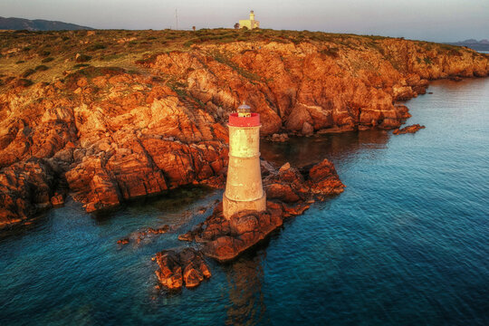 Aerial view of Capo Ferro lighthouse standing on rocky cliffs surrounded by the Mediterranean Sea during golden hour in Capo Ferro, Sardegna, Italy.