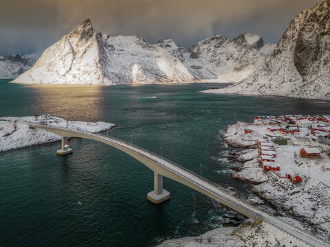 Aerial view of the iconic Hamnoy bridge connecting red rorbu cabins on snow-covered islands against a backdrop of dramatic, snow-capped mountains in Hamnoy, Nordland, Norway.
