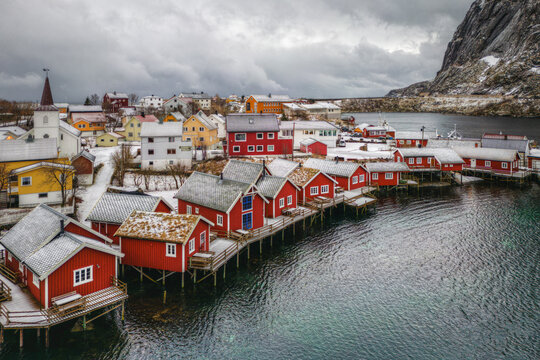 Aerial view of the picturesque village of Reine with its iconic red rorbuer cabins on stilts, snow-dusted rooftops, and dramatic mountain backdrop in Reine, Nordland, Norway.