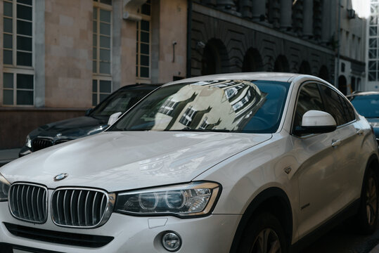 white BMW X4 is parked along a city street. The vehicle reflects the architecture of surrounding buildings on its windows