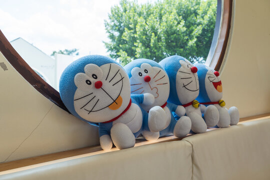 Nakhon Pathom, Thailand - 3 Sep 2026: Four Doraemon plush toys lined up on a seat by a large circular window inside a modern cafe setting.