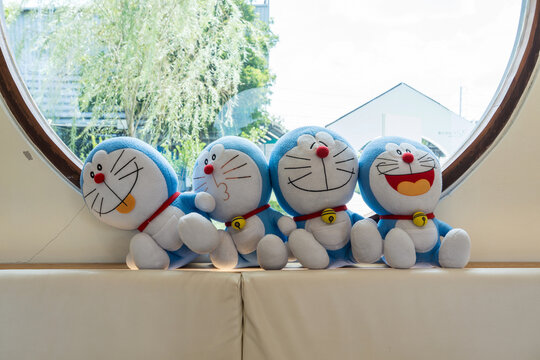 Nakhon Pathom, Thailand - 3 Sep 2026: Four Doraemon plush toys lined up on a seat by a large circular window inside a modern cafe setting.