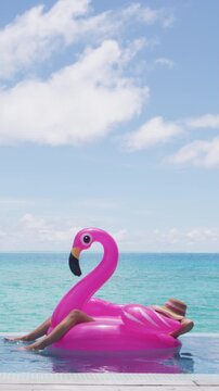Funny travel vacation holidays concept. Woman in pool by beach sunbathing in bikini in inflatable pink flamingo toy float relaxing in luxury. Raw vertical video of real woman model without makeup