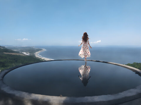The a young woman in a floral dress stands atop a reflective pool overlooking a vast ocean and coastline under a bright sky.