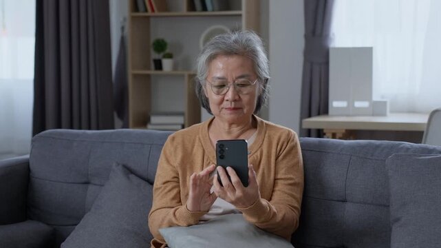 Senior woman looking at smartphone and using mobile application while sitting comfortably on sofa at home, enjoying technology during independent retirement life.