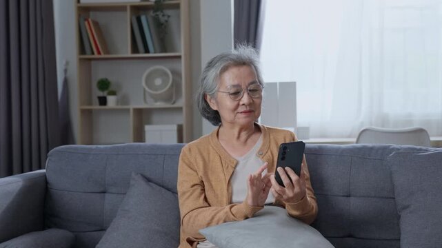 Elderly woman using smartphone and browsing touchscreen while relaxing on sofa at home, exploring mobile technology and digital communication in daily life.