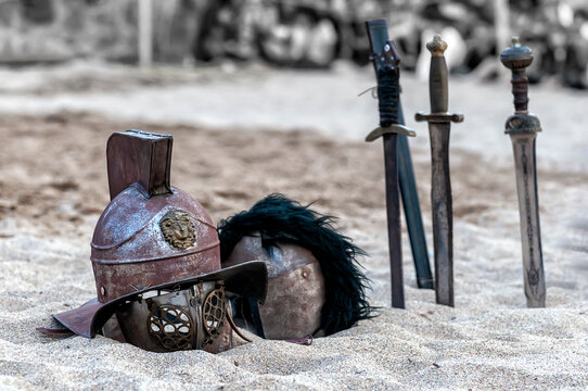 Cascos de gladiador romano con crestas y varias espadas clavadas en suelo arenoso al aire libre durante una recreaci&oacute;n hist&oacute;rica.