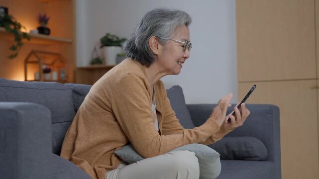Smiling senior woman using smartphone while leaning forward on sofa at home, enjoying mobile technology and staying digitally connected during retirement
