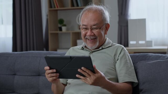 Happy senior man laughing while watching digital tablet on sofa at home, enjoying online entertainment and modern technology during a relaxed retirement lifestyle