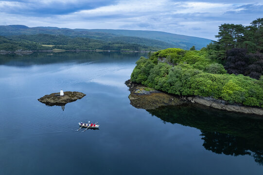 Tighnabruaich, United Kingdom - 28 July 2022: Aerial view of Caladh Harbour with a rowing boat on calm water, surrounded by lush green trees and rocky coastline.