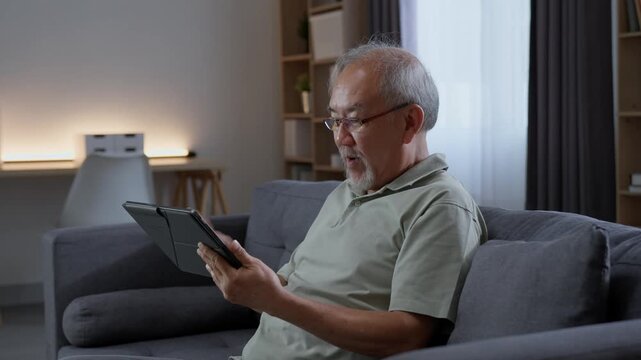 Senior man tapping and browsing digital tablet while sitting on sofa at home, using touchscreen technology to explore online content during his everyday retirement lifestyle.