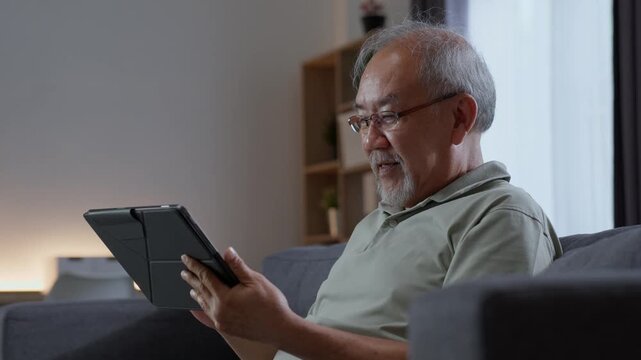 Elderly man concentrating while using digital tablet on sofa at home, reading and browsing online content with modern touchscreen technology during retirement.