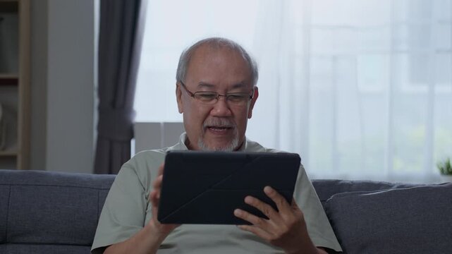 Senior man watching and reading content on digital tablet while relaxing on sofa at home, using modern online technology for information and leisure during retirement