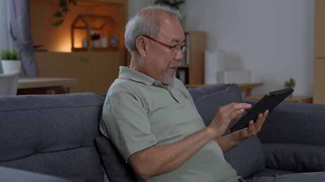Senior man tapping digital tablet screen while sitting comfortably on sofa at home, browsing online content and using touchscreen technology during his retirement leisure time.