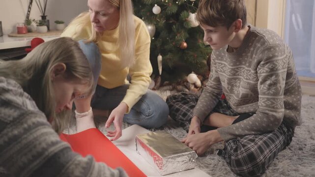 Mother making Christmas memories with son and daughter while wrapping presents beside Christmas tree, sharing traditional holiday activities and joyful family time at home.
