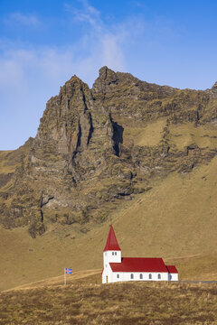 Vik, Myrdalshreppur, Iceland - 10 October 2021: View of the iconic white church with a red roof nestled against the dramatic, rugged mountain slopes under a clear blue sky.