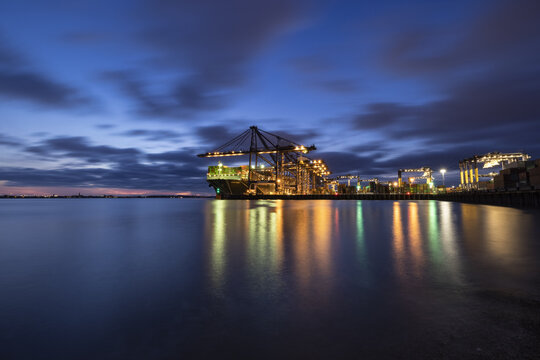 Felixstowe, England, United Kingdom - 26 April 2021: View of Felixstowe Port Container Services at dusk with a cargo ship, illuminated cranes, and reflections on the water under a twilight sky.