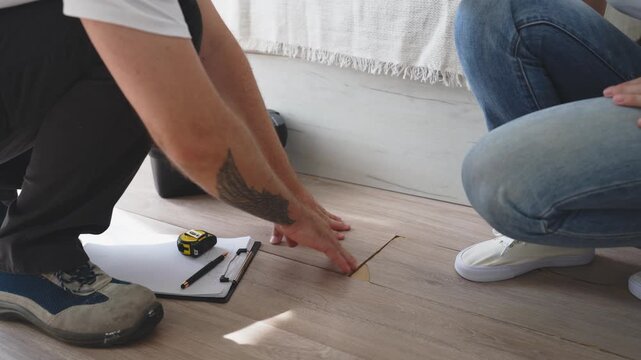 Professional handyman inspecting damaged laminate flooring with female customer, identifying construction defect and discussing home renovation, flooring repair and maintenance options