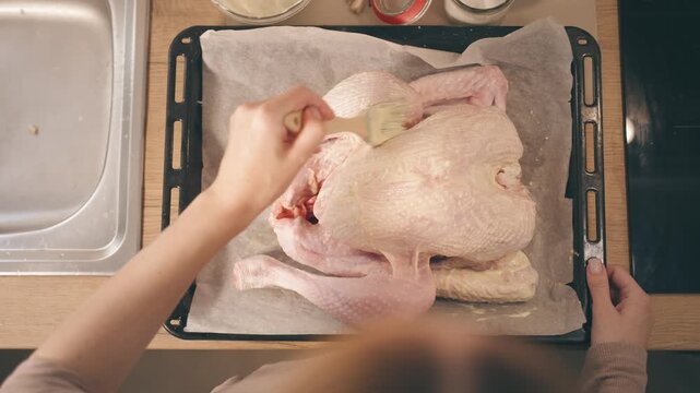 Top-down view of a woman's hands preparing a whole raw turkey for a festive holiday dinner, brushing melted butter over the skin before roasting it for a family meal