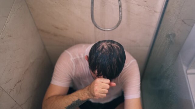Middle-aged man struggling emotionally while sitting alone in shower with wet clothes, representing men's mental health, anxiety, depression and emotional vulnerability.