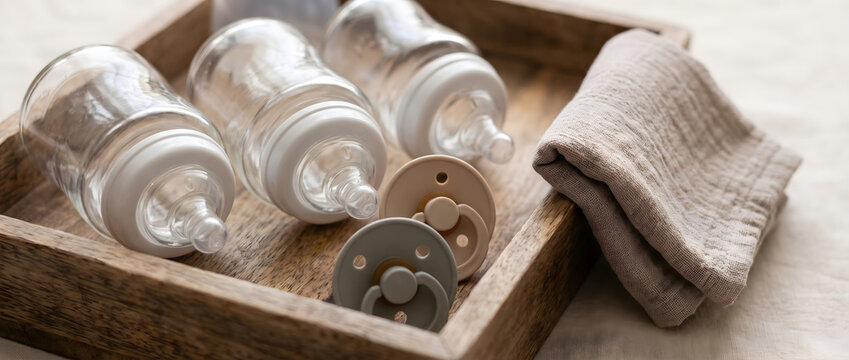 Clear glass baby bottles and pacifiers arranged on a wooden tray, alongside a soft cloth, for infant care.