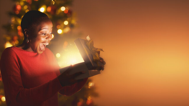 Excited woman gasping with joy opening glowing gift box with blurred Christmas background banner.