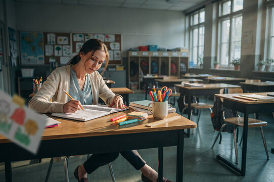 Dedicated female teacher sitting alone at wooden desk grading student notebooks and tests in sunlit empty classroom