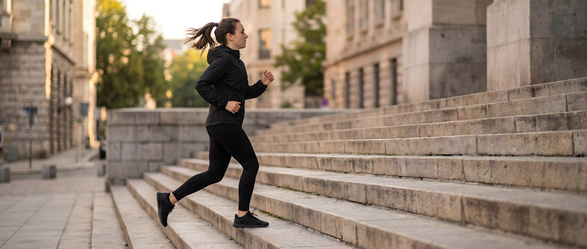 A young woman in black sportswear jogs up stone outdoor stairs in an urban area during daytime.
