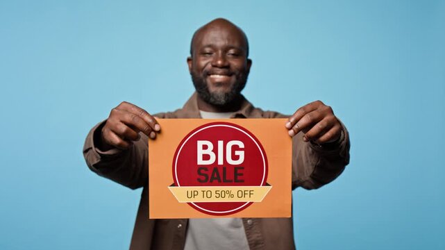 Portrait of young African American man smiling while holding big sale discount sign against blue backdrop. Clip fits retail promotion, seasonal sale advertising, and ecommerce campaigns