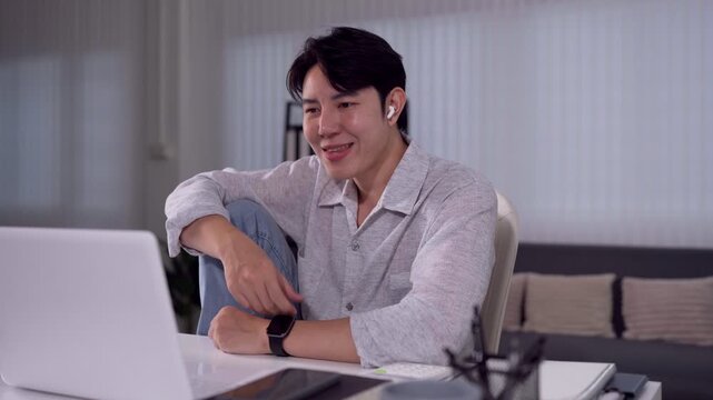 Young Asian man smiling during video call gesturing at laptop while working from home