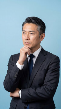 Asian businessman in a suit posing on light blue studio background with his hand on chin, contemplating serious business strategy, facing challenges and developing new ideas for future growth