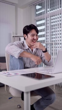 Young Asian man smiling during video call on laptop while working hybrid from modern office desk