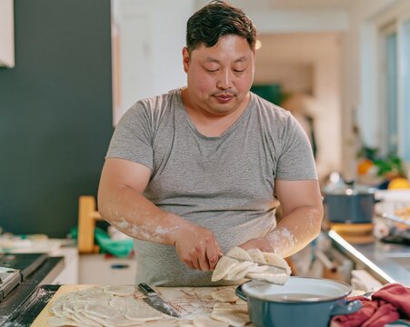 Lunar New Year preparation with a Korean man slicing rice cakes into a blue pot, quiet home cooking with a muted atmospheric mood for culinary and cultural themes