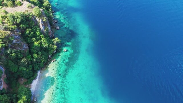 Drone view of a scenic road winding along rocky, forested cliffs and the turquoise waters of Lake Ohrid near Ohrid, North Macedonia. Natural shoreline, secluded beach and clear water on a sunny summer