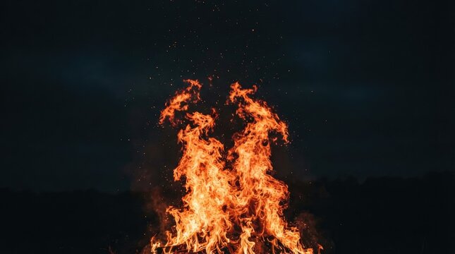 A large bonfire burns intensely against a dark night sky. Flames rise dramatically, creating a dynamic and fiery spectacle.